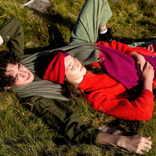 Male and female lying on grass wearing a large scarf in a field, with the scarf showcasing a plain weave design in a purple color.